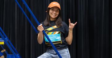 A student stands on a production ladder in an RTF t-shirt and baseball cap with her hand in a "hook 'em" sign.