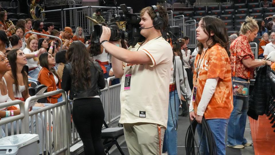 A student wearing over-the-ear headphones holds up a video camera at a sporting event. 
