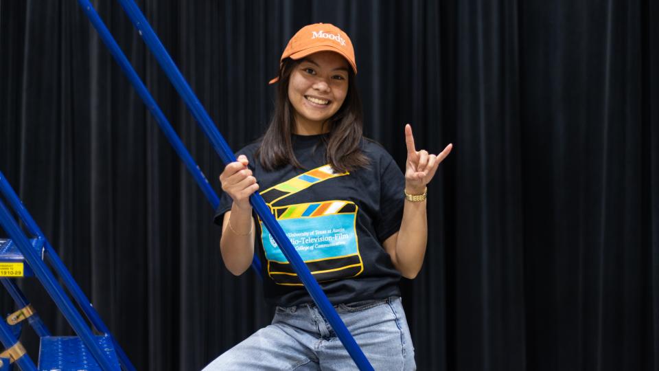 A student stands on a production ladder in an RTF t-shirt and baseball cap with her hand in a "hook 'em" sign.
