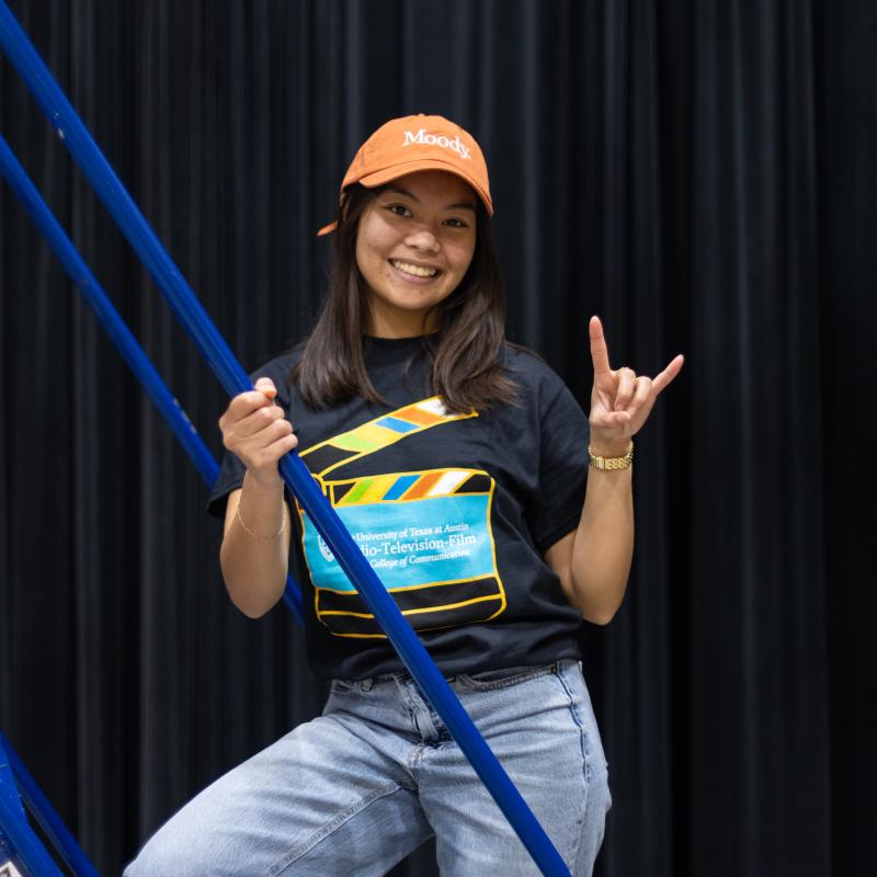 A student stands on a production ladder in an RTF t-shirt and baseball cap with her hand in a "hook 'em" sign.