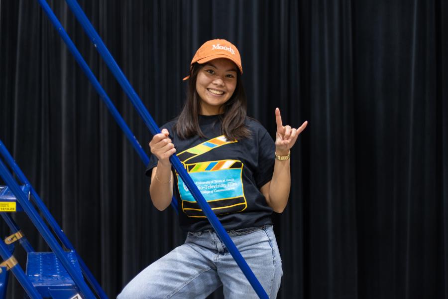 A student stands on a production ladder in an RTF t-shirt and baseball cap with her hand in a "hook 'em" sign.