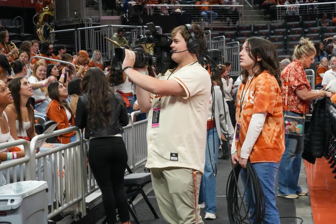 A student wearing over-the-ear headphones holds up a video camera at a sporting event. 